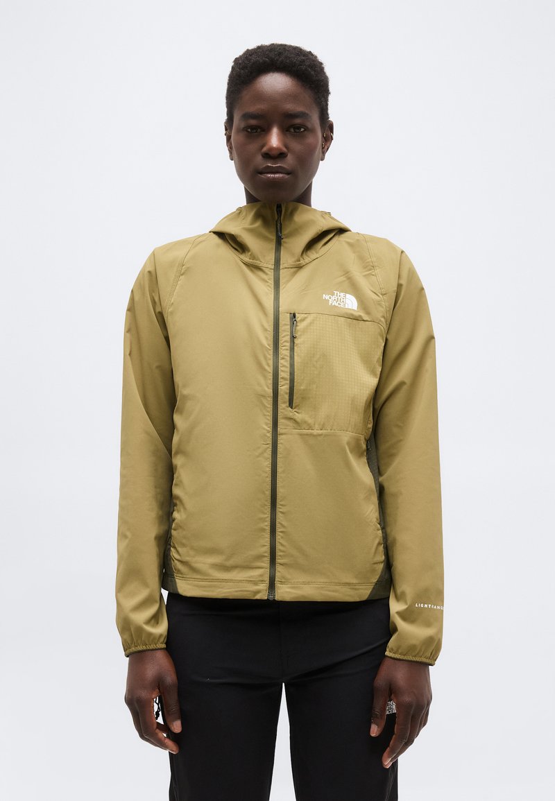Personne portant une veste zippée à capuche The North Face de couleur vert olive, debout devant un fond clair uni.