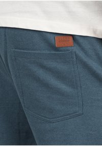 Dark teal sweatpants featuring a patch pocket and a brown branded label. The fabric has a soft texture and a relaxed fit design.