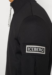 Chaqueta negra con cremallera hecha de tejido texturizado, con un cuello alto y un parche bordado de "ICEBERG" en la parte superior del brazo.