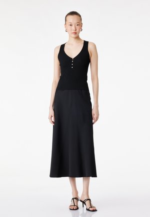 Młoda kobieta ubrana w czarny, bezrękawnik o prążkowanej fakturze z guzikami oraz czarną spódnicę midi, stojąca na białym tle.