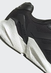 Zapatilla deportiva negra con parte superior de tela texturizada, suela blanca acolchada y detalles en negro. Presenta un diseño elegante y un detalle de logo.