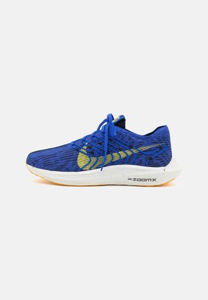 Blue athletic shoe with yellow accents, featuring a mesh upper, rounded toe, and a white midsole with orange outsole. Laced design.
