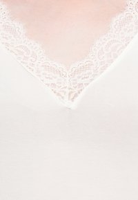 Gros plan sur une poitrine pâle portant un vêtement blanc avec un décolleté en V bordé de délicate dentelle blanche.