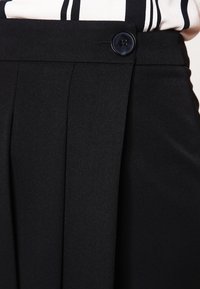 Pantalon noir sur mesure avec plis, présentant une texture lisse, des boutons à la taille et une coupe structurée.