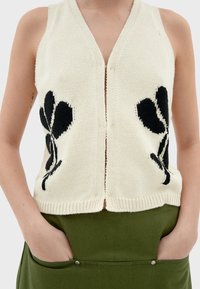 Crème gebreide vest met een V-hals, voorzien van zwarte bloemenpatronen aan beide zijden en een opening aan de voorkant. Gekocht over een groene rok.