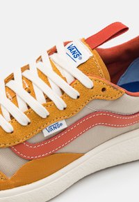 Närbild av orangea, beige och röda Vans-sneakers med vita snörning, sueda- och kanvastexturer, samt synlig varumärkeslogo på tungan och sidan.