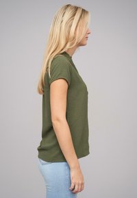 Top vert olive à manches courtes avec une coupe décontractée, en tissu texturé, encolure ronde et ourlet légèrement incurvé. Porté avec un jean bleu clair.