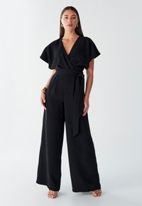 Czarny kombinezon z szerokimi nogawkami, głębokim dekoltem w kształcie litery V, krótkimi falbanowymi rękawami oraz wiązaniem w talii. Gładka tkanina, minimalistyczny design.
