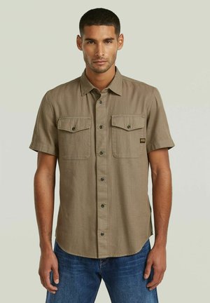 Uomo che indossa una camicia beige a maniche corte con due tasche sul petto, in piedi con le mani rilassate, davanti a uno sfondo semplice.