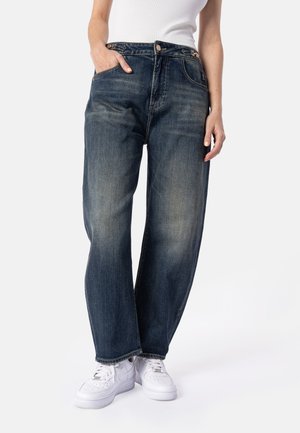 Person iført løse mørkeblå jeans, hvid ærmeløs top og hvide sneakers, med én hånd i lommen, stående mod en ensfarvet baggrund.