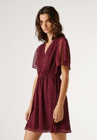 Vestido burdeos con lunares blancos, mangas cortas transparentes, escote en v y cintura elástica. Tela fluida y texturizada con dobladillo por encima de la rodilla.