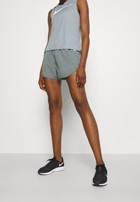 Débardeur gris clair avec logo Nike blanc, associé à un short vert foncé et des chaussures de sport noires avec des accents blancs.