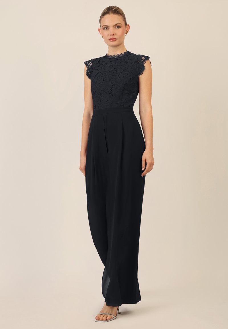 Jumpsuit de encaje negro con un corpino ajustado, mangas en forma de capa y pantalones de pernera ancha con pliegues. Tejido suave con delicados detalles de encaje.