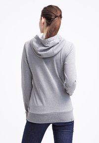 Felpa grigia con una texture morbida, dotata di cappuccio con cordino e orlo a coste. Indossata sopra jeans scuri, mostrando la vista posteriore.