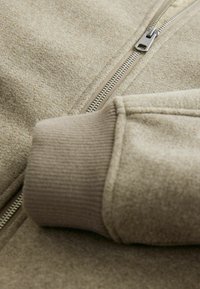Beige sweatshirt med lynlås, der har blødt struktureret stof, ribbede manchetter og en glat metal lynlås detalje.