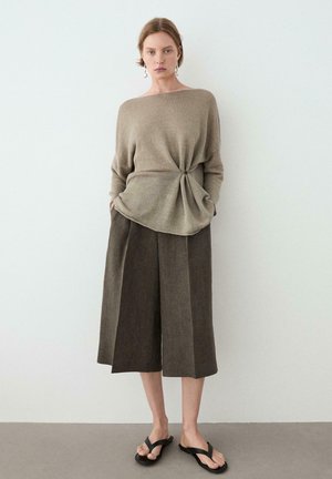 Kobieta w taupe'owym długim dzianinowym swetrze z węzłem z przodu, ciemnobrązowych szerokich, krótkich spodniach, czarnych sandałach i zwisających kolczykach z pereł.