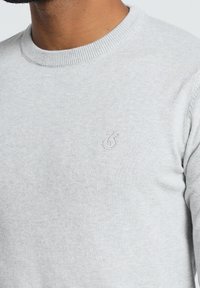 Pull en tricot gris clair avec un col rond, présentant une texture subtilement motifée et un logo brodé sur la poitrine.