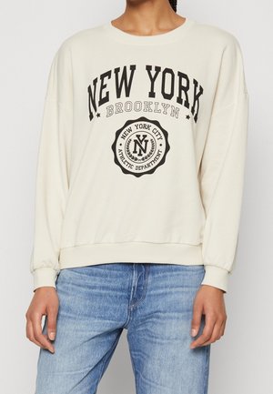 Osoba ubrana w kremową bluzę z napisem "New York Brooklyn" oraz "New York City Athletic Department" w czerni, w połączeniu z niebieskimi dżinsami.