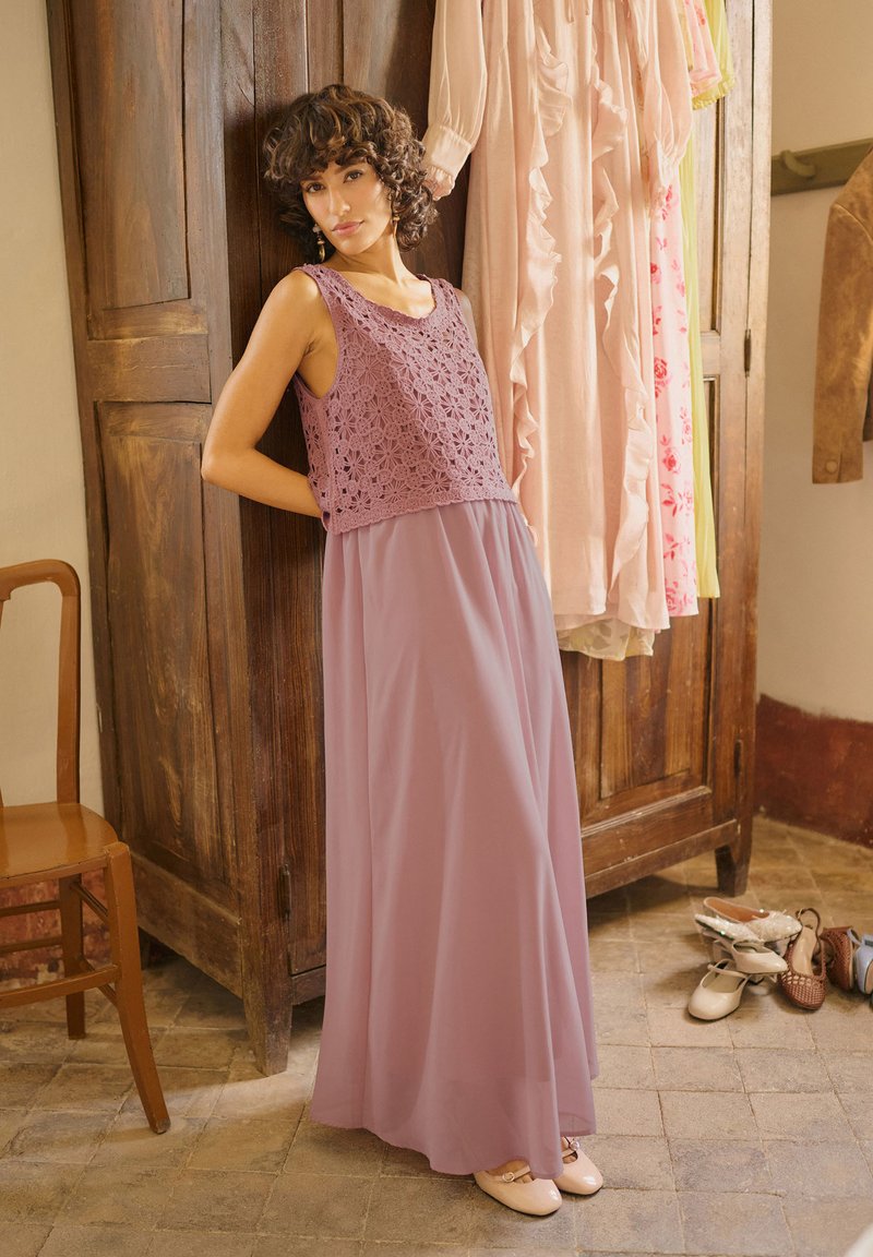 Femme aux cheveux courts et bouclés portant un haut sans manches en dentelle mauve et une longue jupe, appuyée contre une armoire en bois avec des robes et des chaussures à proximité.