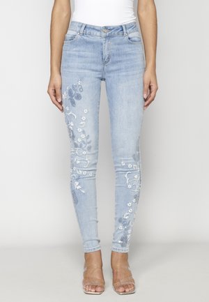 Kvinde iført lyseblå skinny jeans med hvid blomsterbroderi på benene, kombineret med klare remme højhælede sandaler.