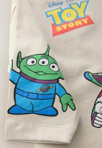 Světle béžové tričko s kresleným šelmem s zelenou pletí, velkýma očima a modrým oblečením, spolu s logem "Toy Story" v žluté a modré.