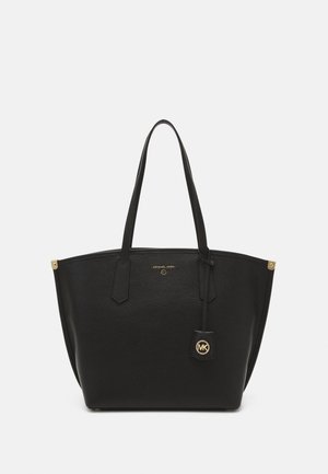 Sac cabas en cuir noir Michael Kors avec double poignées, pendentif logo doré et petits accents dorés aux coins sur un fond neutre.