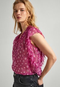 Sheer pink blouse with ruffled sleeves, featuring a diamond pattern and smocked neckline. Paired with dark jeans.
