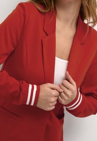 Rød blazer med skræddersyet pasform, hakkraver og hvide stribede accenter på ærmerne. Glat stoftekstur og enkelt knaplukning.