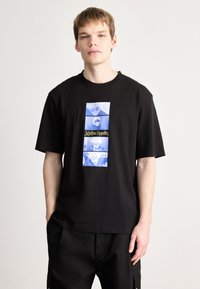 Μαύρο βαμβακερό μπλουζάκι με γραφικό τύπωμα σε μπλε αποχρώσεις και κείμενο σε ορθογώνιο σχέδιο στο μπροστινό μέρος. Κοντά μανίκια και χαλαρή εφαρμογή.