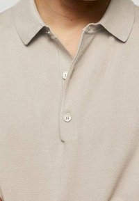 Beige polo paita, joka sisältää klassisen kauluksen ja kolmen napin kiinnityksen. Materiaali on pehmeää neulostekstiiliä, jossa on hienostunut kiilto.