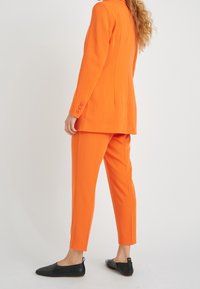 Oranje blazer en bijpassende tapse broek, beiden in een gladde stof. Zwarte, platte, puntige schoenen voltooien de look, met een minimalistisch ontwerp.
