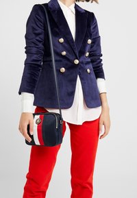 Tummansininen samettinen kaksirivinen bleiseri kultanapeilla, valkoinen napillinen paita, punaiset housut ja tummansininen käsilaukku punaisilla ja valkoisilla raidoilla.