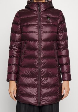 Kobieta ubrana w długą, błyszczącą, ciemnofioletową puchową kurtkę pikowaną z kapturem i zamkiem z przodu, stojąca na jednolitym tle.