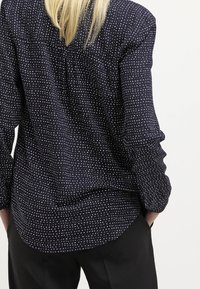 Blusa blu navy con pois bianchi, maniche lunghe e dettaglio sullo yoke posteriore. Tessuto morbido con una vestibilità ampia e orlo curvo.