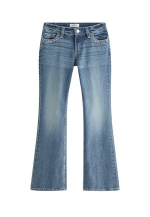 Teenagepige står iført en hvid kortærmet skjorte, blå udvidede jeans og hvide sneakers mod en ensfarvet lys baggrund.