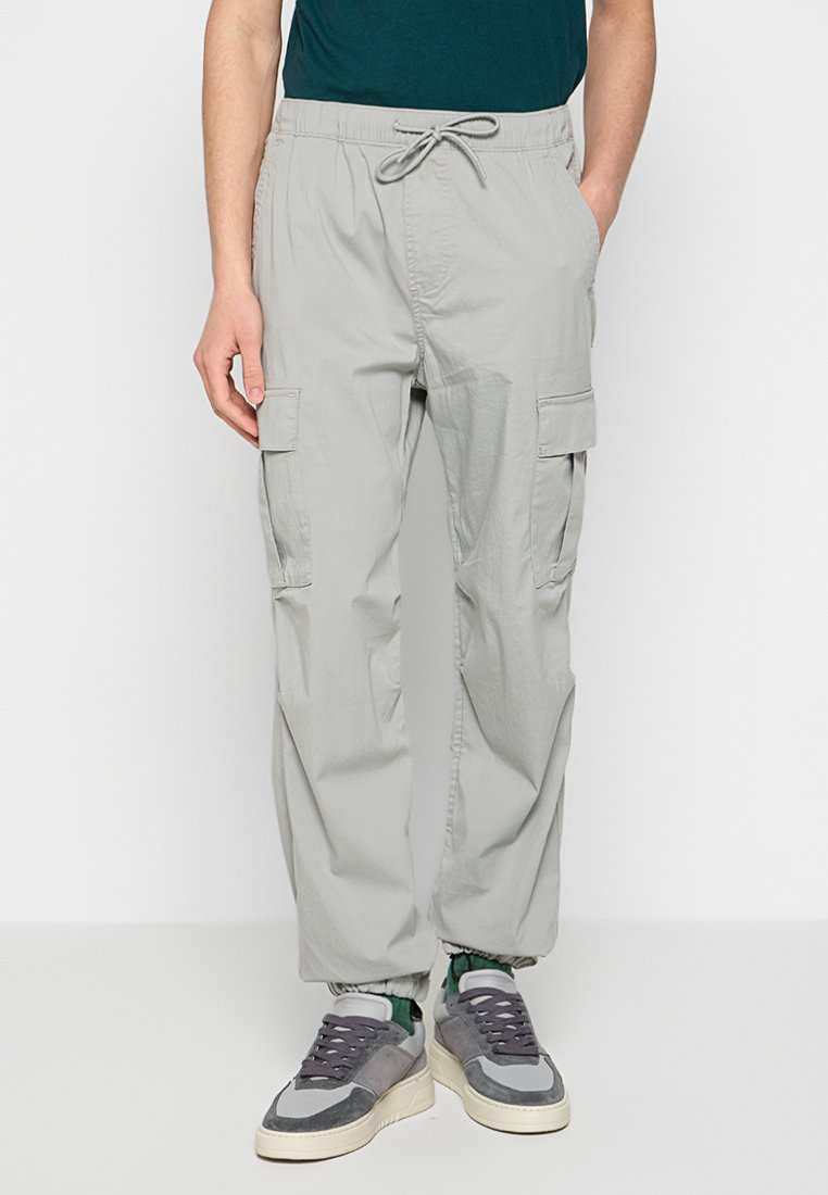 Helle graue Cargo-Hose mit elastischem Bund und Kordelzug, ausgestattet mit Seitentaschen und lockerer Passform, kombiniert mit lässigen grauen Sneakers.