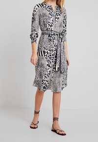 Leopardenprint jurk in zwart en wit met een ceintuur om de taille, een knoopsluiting aan de voorkant en lange mouwen. Bevat verschillende textuurpatronen en strepen.