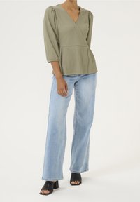 Olivgrüne, strukturierte Bluse mit V-Ausschnitt und dreiviertel Puffärmeln, kombiniert mit hellblauen, weit geschnittenen Jeans und schwarzen Sandalen mit Absatz.