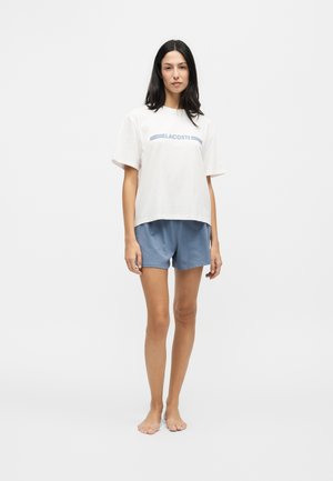 Femme debout pieds nus portant un t-shirt blanc Lacoste et un short bleu sur un fond gris clair uni.