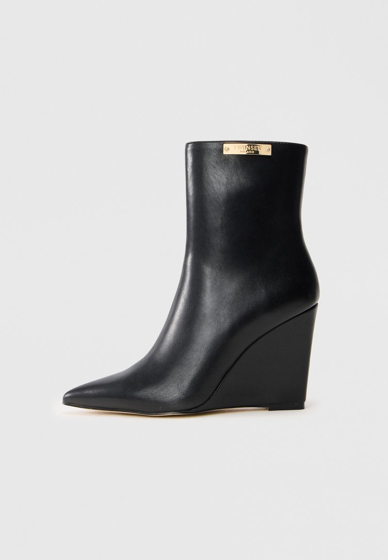 Bottine à talon compensé en cuir noir avec un bout pointu et une plaque de marque dorée près du bord supérieur, présentée sur un fond blanc.