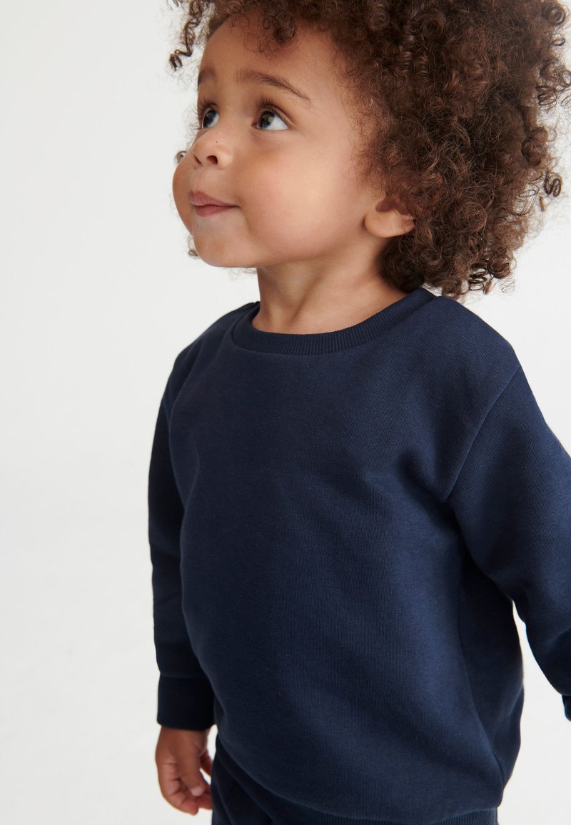 Junges Kind mit lockigem Haar, das ein dunkelblaues Langarmshirt trägt und leicht nach oben links auf einem hellen Hintergrund schaut.