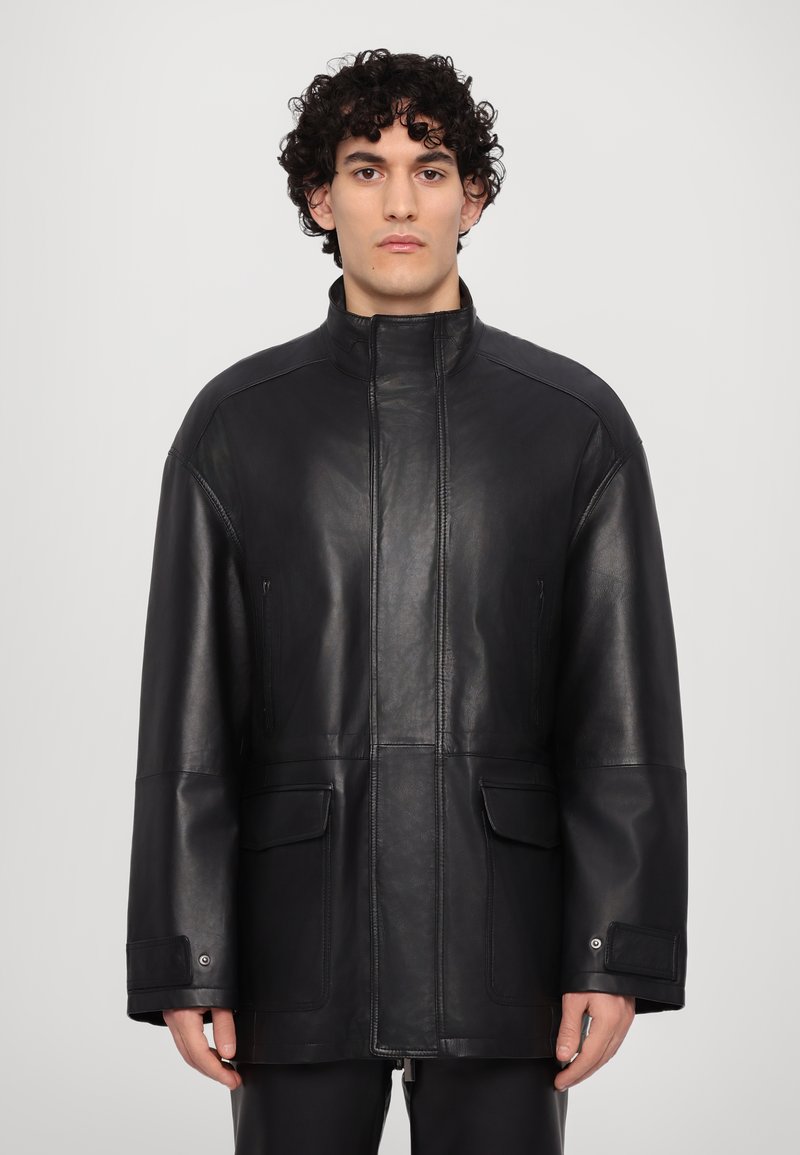 Chaqueta de cuero negra con un cuello alto, dos bolsillos frontales y un corte holgado. Textura suave con detalles de costura mínimos.