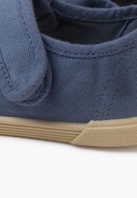Zapato de tela azul con un empeine texturizado, que presenta una punta redondeada y una suela de goma beige. Incluye una lengüeta acolchada y detalles cosidos.