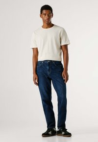 Camiseta de algodón beige de manga corta, combinada con jeans de denim azul oscuro. Modelo vistiendo zapatillas negras con acentos blancos.