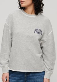 Sudadera gris claro con mangas largas, cuello redondo acanalado y logo bordado "Superdry Athletics Beach Club" en azul marino. Textura de tejido suave.