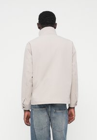 Veste gris clair à col montant, avec une texture lisse et des manches longues, associée à un jean en denim bleu. Design minimaliste.