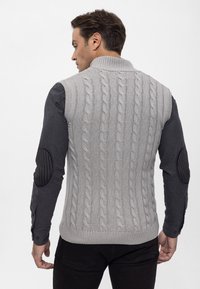 Grau gestrickte Weste mit einem gerippten Kragen, kombiniert mit dunklen langen Ärmeln, die strukturierte schwarze Ellbogenpatches und eine taillierte Silhouette aufweisen.