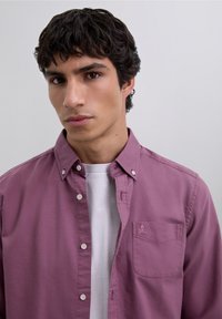 Lila Hemd zum Knöpfen mit langen Ärmeln und einer Brusttasche, die ein kleines Totenkopf-Logo zeigt. Wird über einem weißen T-Shirt getragen. Glatte Textur.