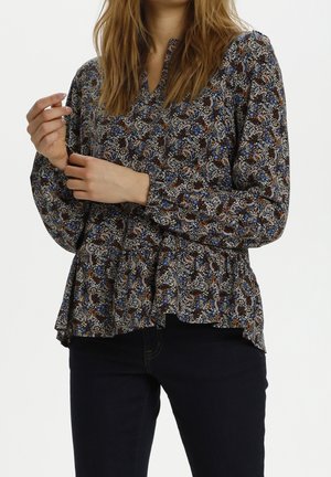 Vrouw die een bloes met lange mouwen en een bloemenpatroon draagt, met een peplumzoom en donkere broek, haar linker mouw verstelend.