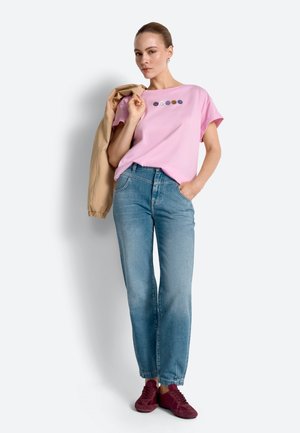 Žena stojaca s béžovým sakom cez rameno, oblečená v ružovom tričku s ikonami usmievavých tvárí, modrých džínsoch a marónových teniskách na bielom pozadí.
